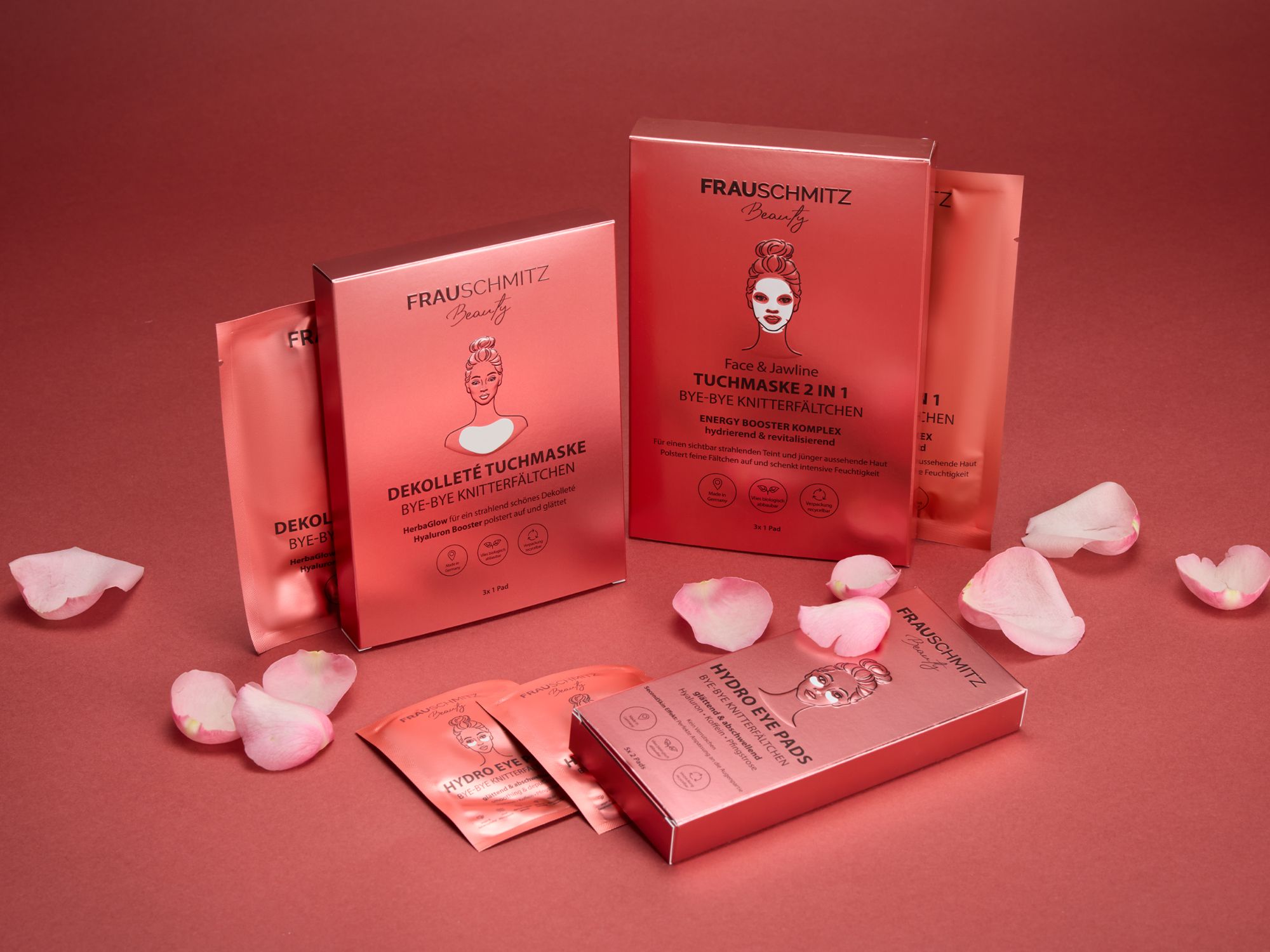 Mehrere rosa Verpackungen von FRAUSCHMITZ Beauty Produkten, darunter Dekolleté Tuchmaske und Face & Jawline Tuchmaske 2 in 1, umgeben von rosa Blütenblättern.