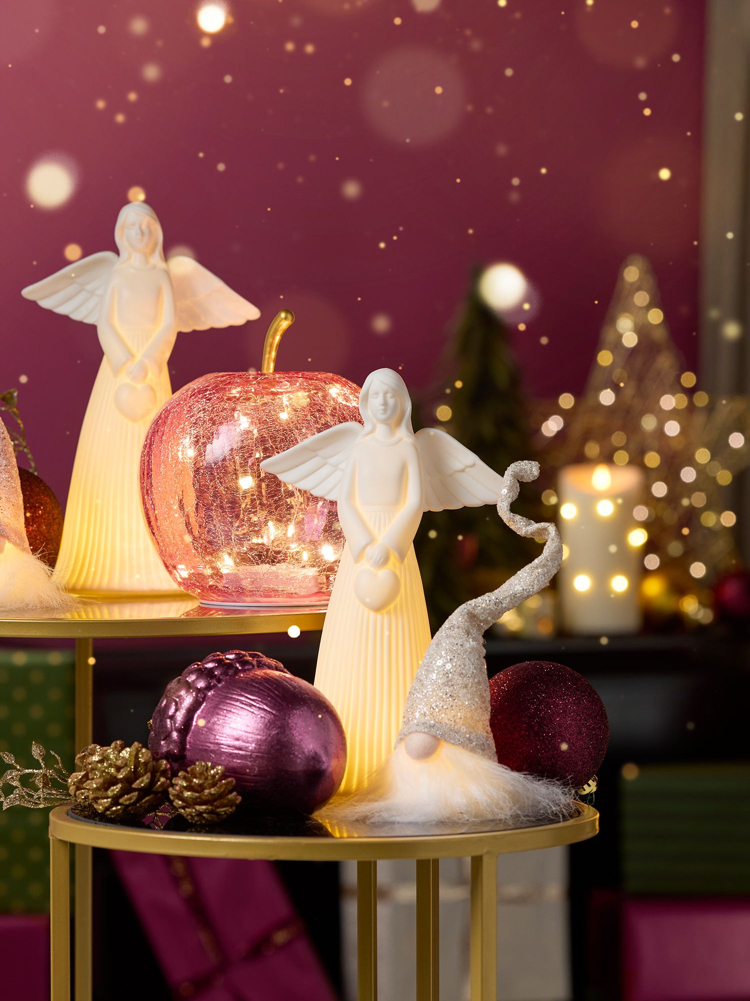 Weihnachtliche Wohnideen Zwei weiße Engel-Figuren mit Flügeln auf goldfarbenem Tisch, umgeben von glitzernden Weihnachtskugeln, Zapfen und Lichterketten.