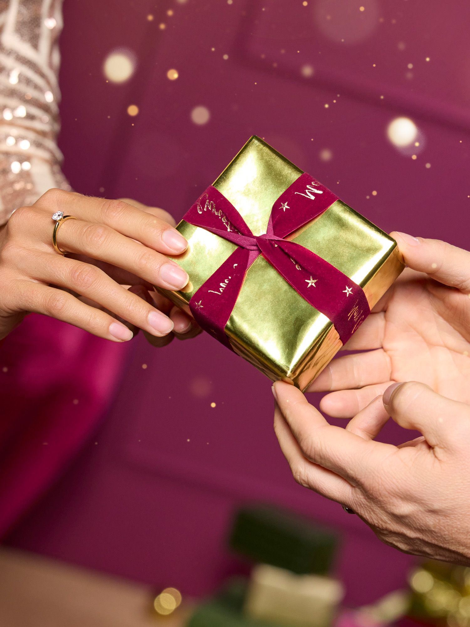 Geschenkideen unter 25€ Zwei Hände übergeben ein kleines Geschenk in goldener Folie mit rotem Band, auf dem Sterne und Schrift zu sehen sind.