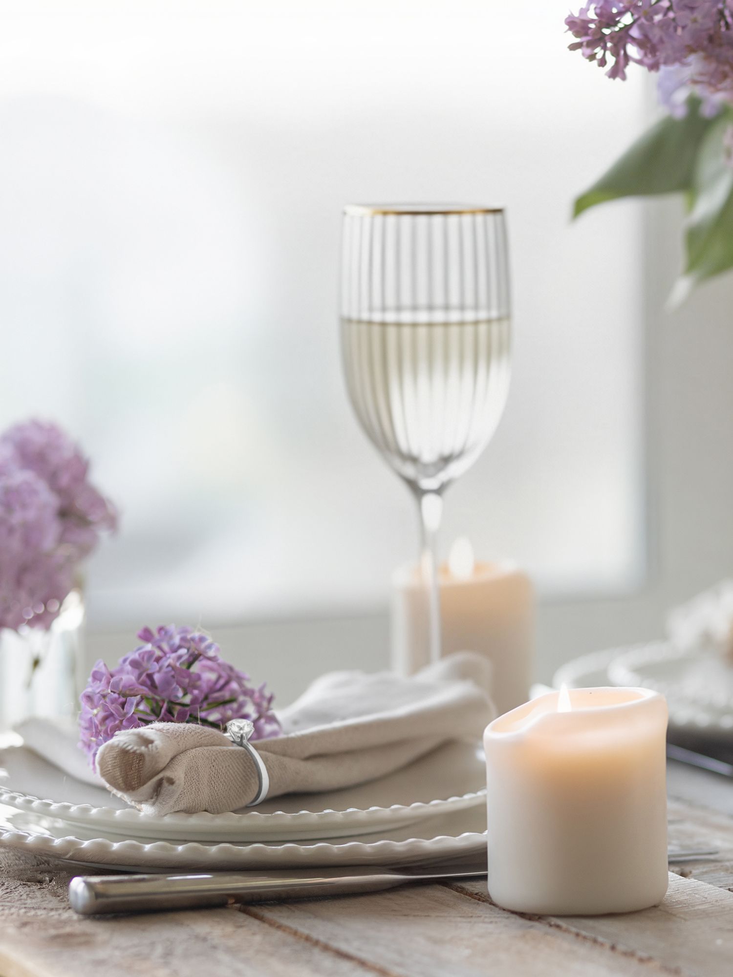 Gedeckter Holztisch mit weißen Kerzen, goldrandigen Weingläsern, weißen Tellern, Stoffservietten und lila Fliederblüten