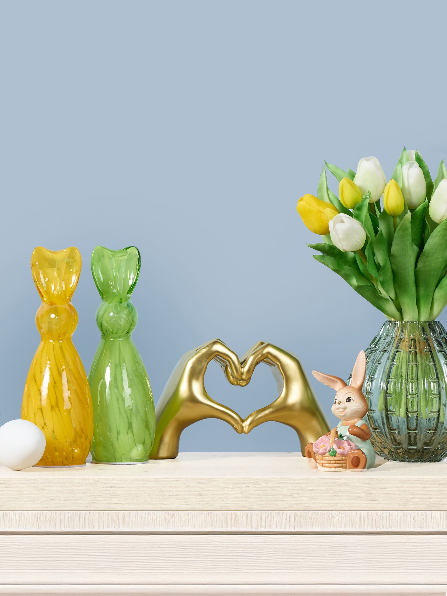 Dekoration Gelbe und grüne Hasenfiguren aus Glas, goldene Hände, die ein Herz formen, weiße Eier in Schalen und ein Glasvase mit gelben und weißen Tulpen vor blauem Hintergrund
