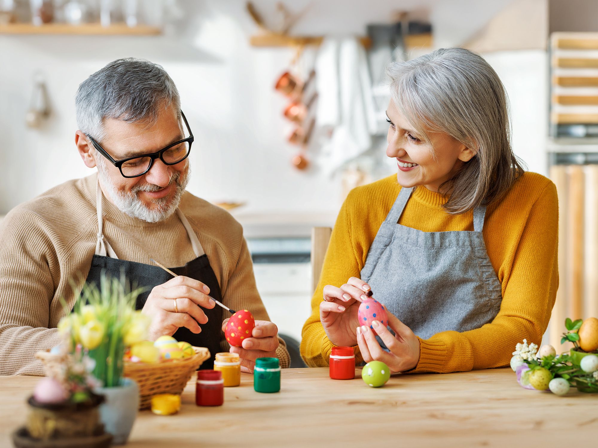 Zwei ältere Personen sitzen an einem Tisch und bemalen Ostereier, umgeben von Osterdekoration und Farbtöpfen