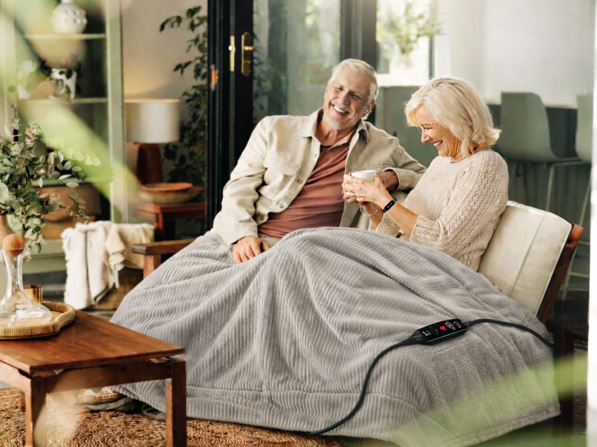 Älteres Paar sitzt auf Sofa unter einer grauen, gerippten Heizdecke mit Fernbedienung
