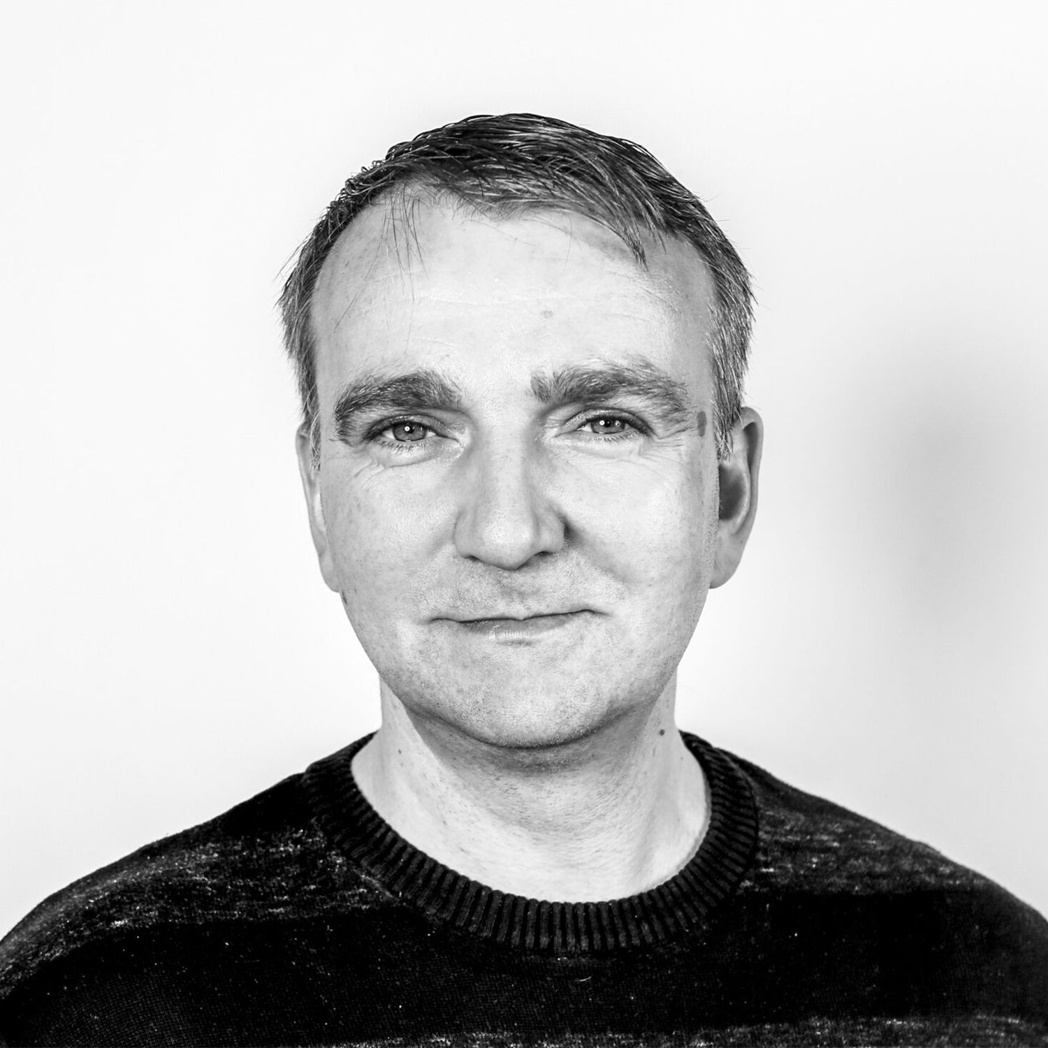 Mann mit kurzem Haar trägt einen dunklen Pullover mit Rundhalsausschnitt vor weißem Hintergrund