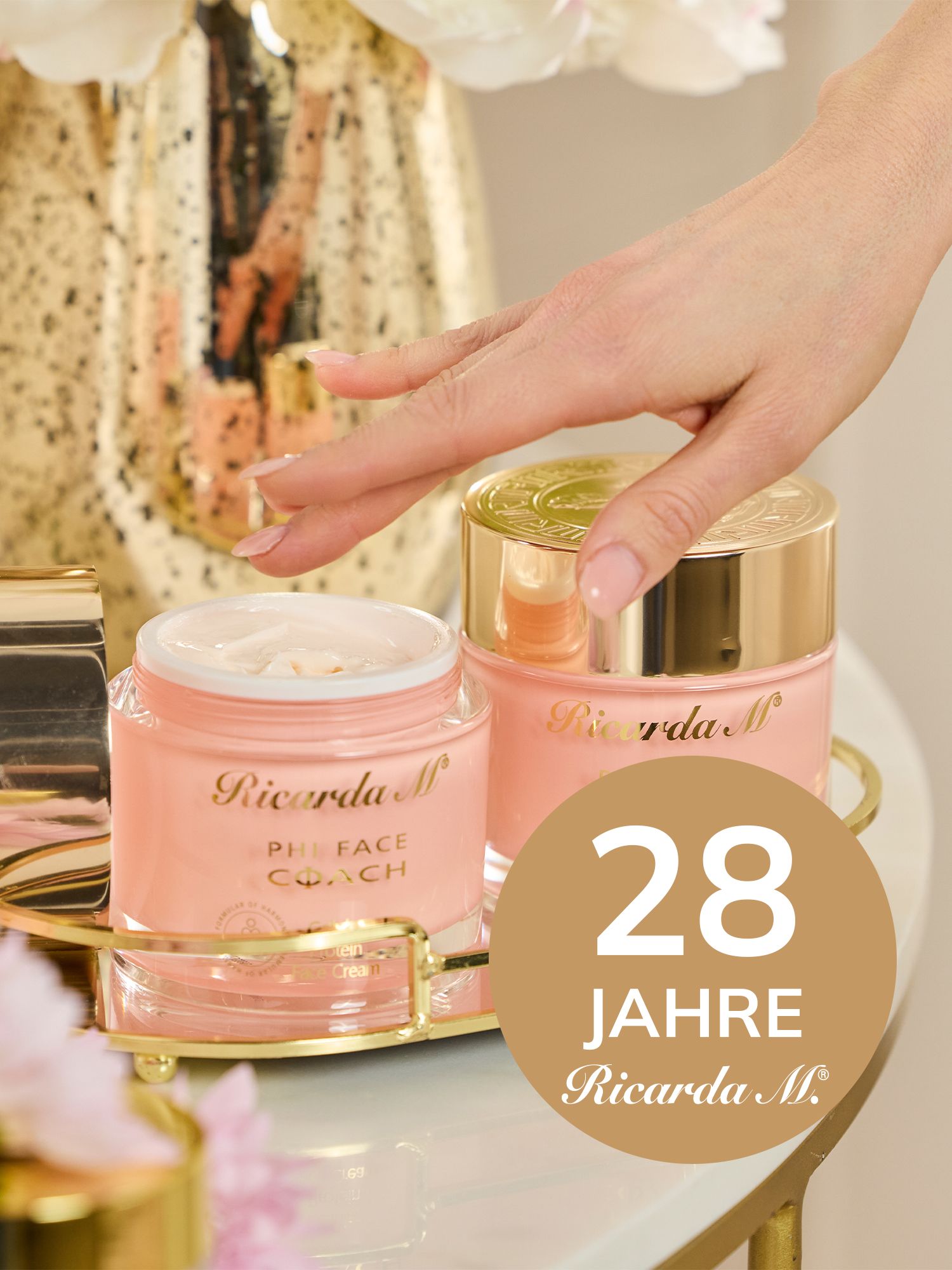 PHI FACE COACH – Entdecken Sie die innovative Hautpflegelinie von Ricarda M! Hand greift nach einem geöffneten, rosafarbenen Gesichtscremetiegel mit goldfarbenem Deckel auf einem goldfarbenen Tablett neben weiteren Kosmetikprodukten